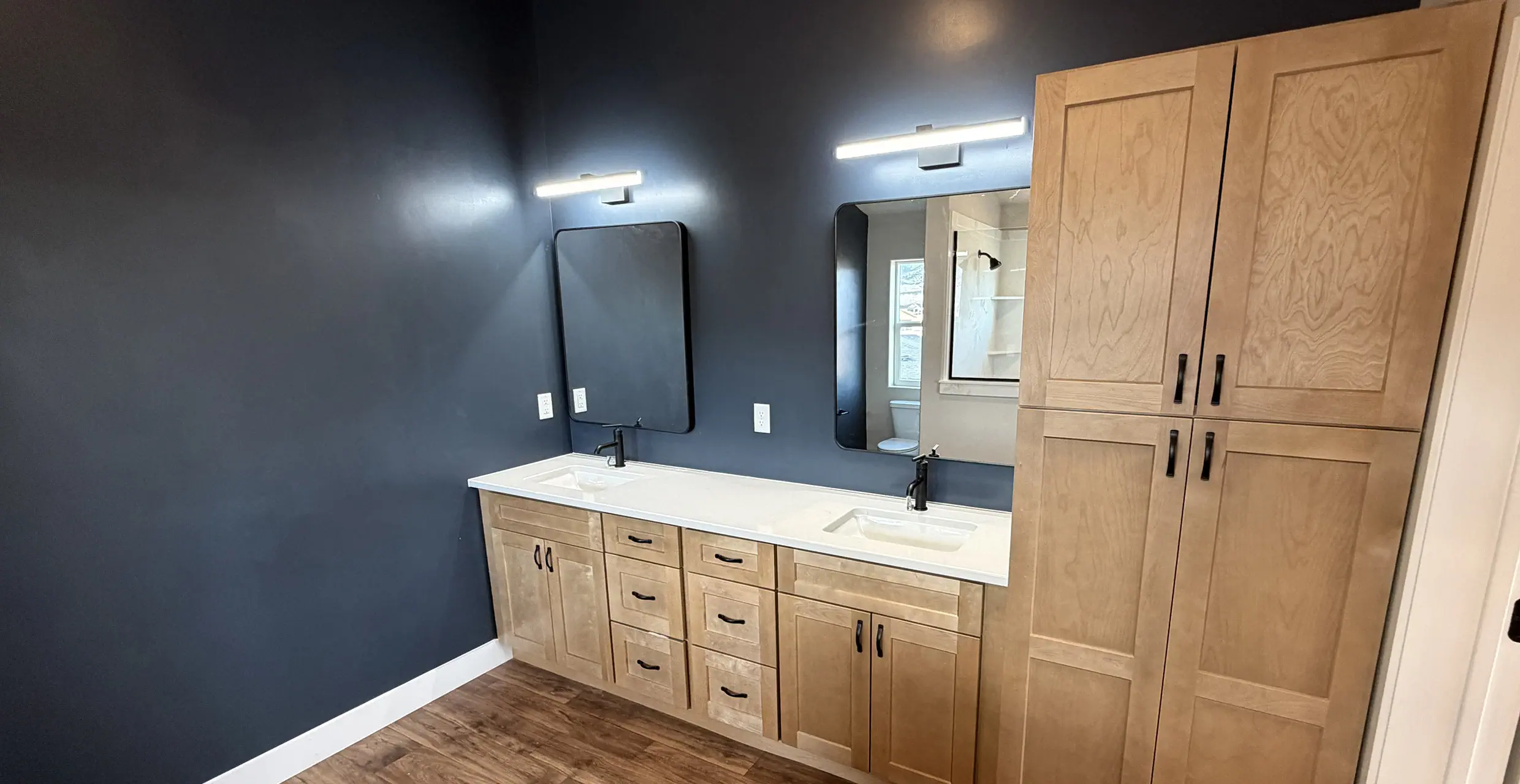 Primary bathroom with navy walls, a maple double vanity, black-framed mirrors and a tall maple linen cabinet in a finished AJ Caplin home